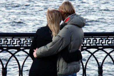 Couple standing by a waterfront railing, embracing while looking out over the water.