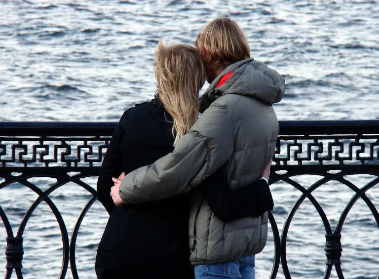 Couple standing by a waterfront railing, embracing while looking out over the water.