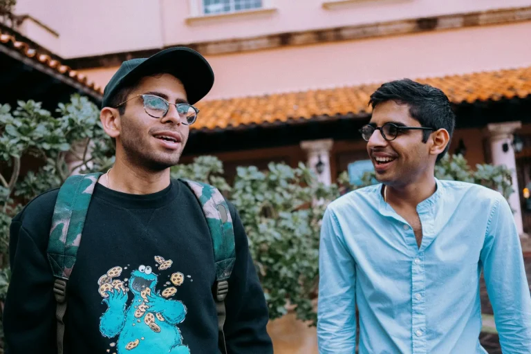 Two young men smiling and talking outdoors near a building with a tiled roof.